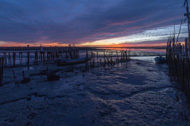 Dalgalar, tarihi ahşap iskeleye yanaşmış çamurlu düzlükleri ve balıkçı teknelerini gösteriyor.