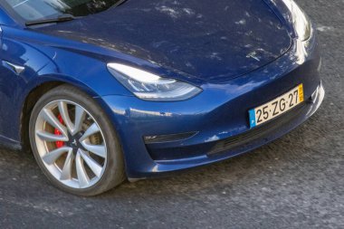 Mavi Tesla modeli 3 elektrikli araç Portekiz plakalı asfalt yolda ilerliyor.