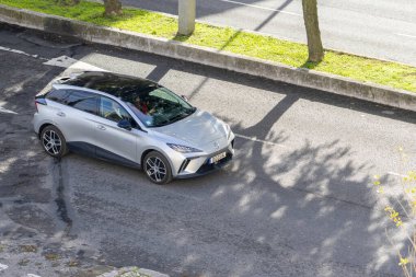 Gümüş MG4 elektrikli hatchback, ağaç gölgeleriyle asfalt bir yolda ilerliyor.