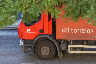 Kırmızı Ctt Correios kamyonu yeşil ağaç dallarının altında asfalt yolda gidiyor.