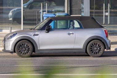 Gümüş rengi mini Cooper. Üstü açık bir araba. Şehir caddesinde bir binanın yanına park edilmiş.