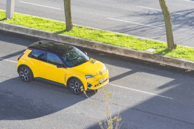 Sarı elektrikli Renault 5 e-teknik hatchback asfalt yolda kaldırımın yanında sürüyor.