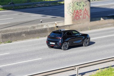 Siyah kompakt araba, çok şeritli bir otoyolda beton sütun kaplı bir duvar yazısının yanında seyahat ediyor.