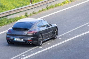 Siyah Porsche Panamera yeşil bitkilerle dolu bir yolda gidiyor.