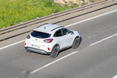 Yol kenarında yeşil bitki örtüsüyle asfalt yolda giden beyaz Ford puma SUv.