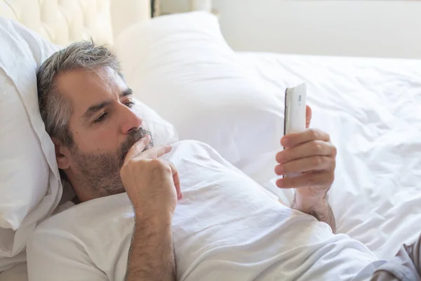 Kıdemli adam düşünceli bir şekilde cep telefonuna bakıyor. Adam yatağa uzanıyor ve cep telefonu kullanıyor..