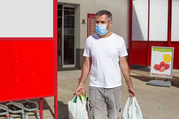 A man in a medical mask leaves the market with full bags of groceries in his hands. A masked man made purchases during the pandemic. Shopping during a pandemic.