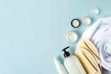 Açık mavi arka planda beyaz banyo havlusu, krem ve banyo terlikleri var. Metin için boş alan.