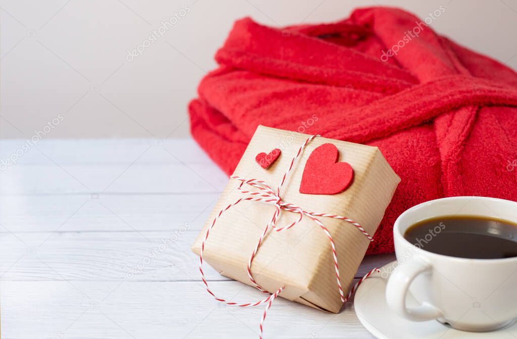 A gift decorated with red hearts for Valentine's Day, a cup with black coffee and a red bathrobe are on a light background. The 14th of February. Free space for text.