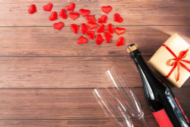 Bir şişe şampanya, iki cam bardak, kırmızı kurdeleli bir hediye ve şişeden çıkan kalpler ahşap arka planda. Sevgililer Günü teması. Metin için boş alan.