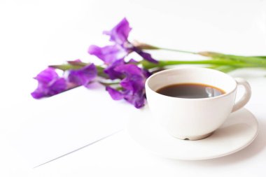 Sade kahve, bir buket iris çiçeği ve açık arkaplanda boş bir kağıt zarf olan beyaz bir fincan. Metin için boş alan.