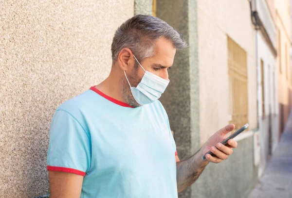 A man in a medical mask and in summer clothes is standing in the street during the day and looking at the mobile phone in his hand.