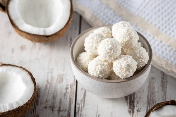 Portions of white chocolate coconut candy balls in the bowl, raw cracked coconut on the background