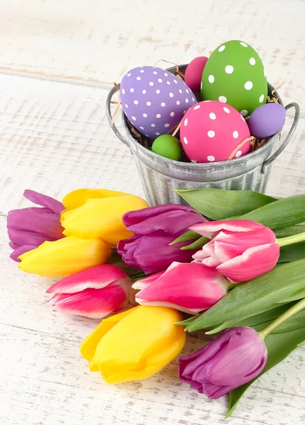Ovos de Páscoa brilhantes em um balde e tulipas de primavera em um contexto de madeira. Um fundo de Páscoa com espaço para o texto . — Fotografia de Stock