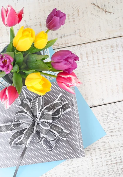 Boeket van heldere lente tulpen en de doos van de gift met een strik op een lichte houten achtergrond. Een feestelijke achtergrond met ruimte voor de tekst. Bovenaanzicht. — Stockfoto