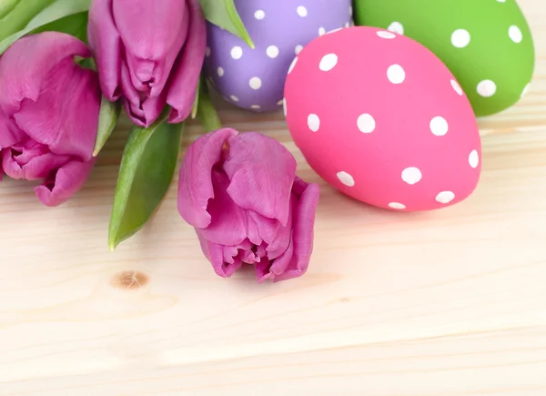 Violet tulips and bright Easter eggs on a light wooden background. An easter background with space for the text. — Stock Photo, Image