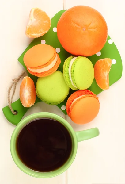 Orange and green macaroons, cup of black tea and tangerine on a white wooden background. Tasty breakfast. Top view. — Stock Photo, Image