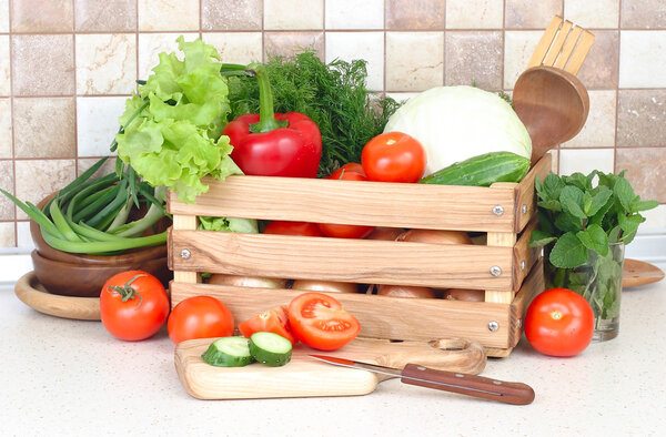 The fresh washed-up vegetables in a wooden box and the cut vegetables on a chopping board against modern kitchen.