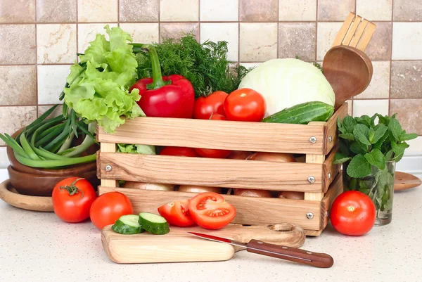 Las verduras frescas lavadas en la caja de madera y hortalizas cortadas en la tabla que corta contra la cocina moderna . — Foto de Stock