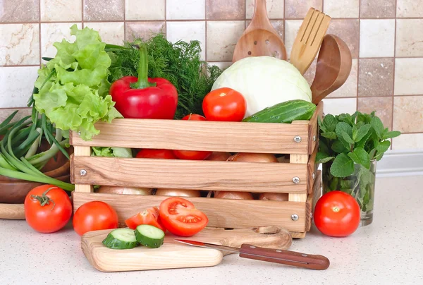 The fresh washed-up vegetables in a wooden box and the cut vegetables on a chopping board against modern kitchen. — Stock Photo, Image