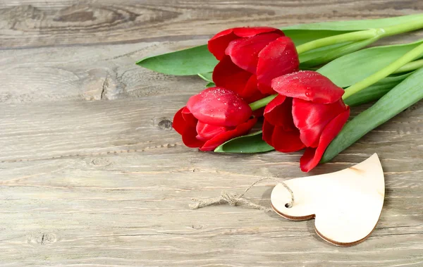 Bouquet of fresh red tulips and wooden heart (a place for the text) on a wooden background. — Stock Photo, Image