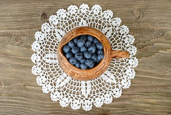 Fresh blueberry in a clay small jug on a lacy napkin on a wooden background. Top view. — Stock Photo, Image
