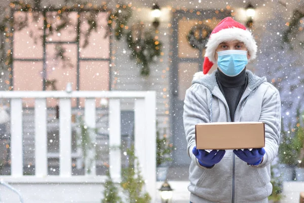 Safe delivery of gifts and packages for the holidays in quarantine during the coronavirus pandemic. A courier, a volunteer in a Santa hat, a medical mask and gloves, holds a box.