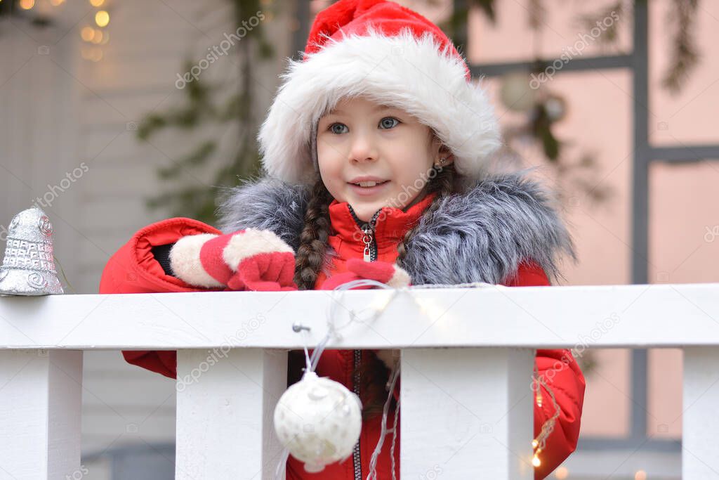 A little girl in a Santa hat with a smile on her face is standing on the porch of a Christmas house. Happy New Year and Merry Christmas!