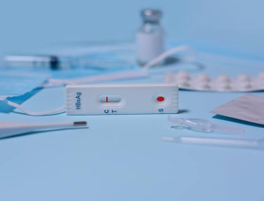 Hepatit B Virüs Testi (HBsAg) için hızlı test kaseti, tıbbi maske, şırınga, termometre, mavi zemin üzerinde ilaç. Test negatif sonuç veriyor. Boşluğu kopyala