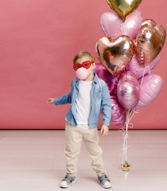 Çocuk dans ediyor, gülümsüyor ve pembe kalp şeklindeki balonların yanında eğleniyor. Pembe arka planda izole edilmiş.