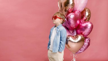 Çocuk dans ediyor, gülümsüyor ve pembe kalp şeklindeki balonların yanında eğleniyor. Pembe arka planda izole edilmiş.