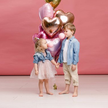 Küçük oğlan ve kız sarılıyor, dans ediyor, gülümsüyor ve pembe kalp şeklindeki balonların büyük dalında eğleniyorlar. Valentins Günü konsepti. Pembe arka planda izole edilmiş.