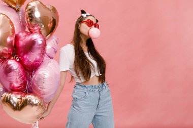 Dans eden, gülümseyen ve sakızla eğlenen genç bir kadın pembe kalp şeklindeki balonların yanında. Pembe arka planda izole edilmiş.