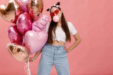 Dans eden, gülümseyen ve sakızla eğlenen genç bir kadın pembe kalp şeklindeki balonların yanında. Pembe arka planda izole edilmiş.