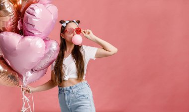 Dans eden, gülümseyen ve sakızla eğlenen genç bir kadın pembe kalp şeklindeki balonların yanında. Pembe arka planda izole edilmiş.
