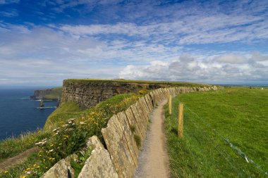 İrlanda 'daki Moher Kayalıkları' nın kenarında mavi gökyüzü, beyaz bulutlar ve arka planda deniz olan bir yol manzarası..
