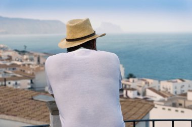 Sırtı dönük ve şapkalı bir adam arka planda deniz olan bir kıyı kasabasının manzarasına bakıyor..