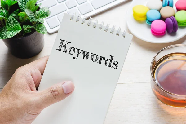 Man holding Keywords message on book and keyboard with a hot cup of tea, macaroon on the table. Can be attributed to your ad. — Stock Photo, Image