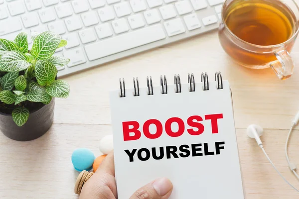 Man holding BOOST YOURSELF message on book and keyboard with a hot cup of tea, macaroon on the table. Pode ser atribuído ao seu anúncio . — Fotografia de Stock