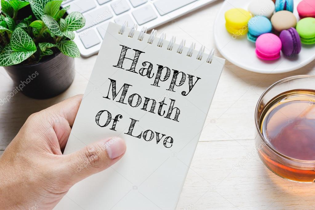 Man holding Happy month of love message on book and keyboard with a hot cup of tea, macaroon on the table. Can be attributed to your ad.