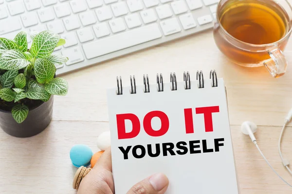 Mann hält Do-it-yourself-Nachricht auf Buch und Tastatur mit einer heißen Tasse Tee, Makronen auf dem Tisch. kann Ihrer Anzeige zugeordnet werden. — Stockfoto