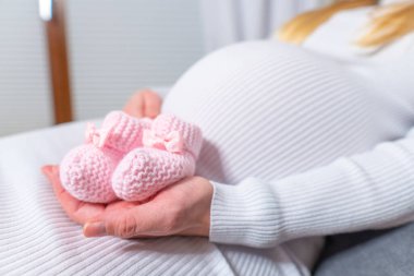 Hamile bir kadının karnındaki doğmamış bebek için küçük ayakkabılar. Küçük bebek ayakkabıları tutan hamile bir kadın yatak odasında dinleniyor. Hamile bir kadının karnındaki doğmamış bebek için küçük ayakkabılar.