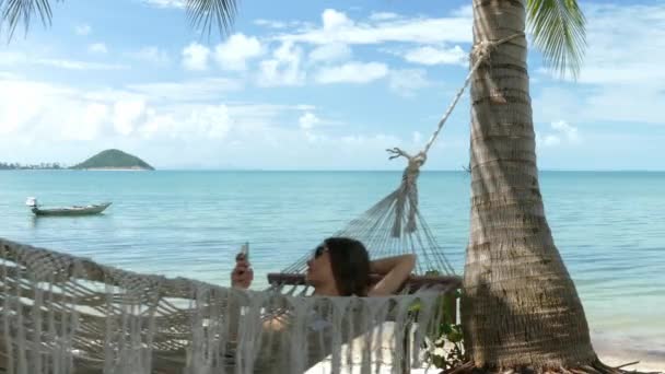 Woman using mobile Kphone in a hammock on the beach — Stock Video
