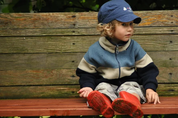 Junge in roten Gummistiefeln auf einer Schaukelbank sitzend. Verschlungene Reben schwingen. — Stockfoto