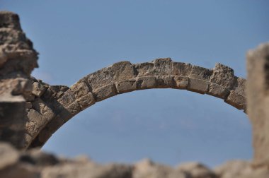 Aşınmış bir taş kemerin kalıntıları arkeolojik bir alanda yerden yükseliyor. Altında antik yapıların parçaları görünüyor ve yukarıda parlak mavi bir gökyüzü var..