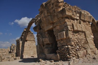 Antik bir taş yapının kalıntıları parlak mavi bir gökyüzüne karşı görülebilir. Yıpranmış bloklar ve kemerler sitenin tarihsel önemini ve ihtişamını gözler önüne seriyor.