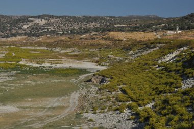 Kuru bir manzara, yeşil çalı ve kayalık arazili geniş bir göl yatağını gösteriyor. Bu sakin ortamda etrafındaki dağlar berrak mavi bir gökyüzünün altında görülebilir..