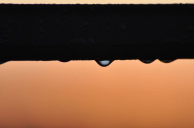 Güneş arka planda batarken bir su damlası korkuluklara tutunur ve gökyüzünde sıcak renkler bırakır. Bir sükunet ve doğanın güzelliğini yakalar..