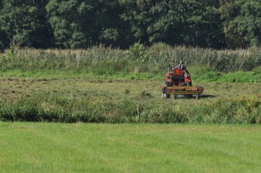 Kırsal kesimde parlak bir gün, büyük, açık bir alanda çim biçen bir traktörü gösteriyor. Yeşillik, arka planda gölgeler oluşturan ağaçlarla birlikte sahneyi çevrelemektedir..
