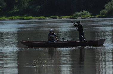 İki birey sakin bir nehirde küçük bir tekneyi yüzdürmek için birlikte çalışıyorlar. Bereketli yeşillik berrak gökyüzünün altındaki suya yansıyarak barışçıl bir atmosfer yaratıyor..